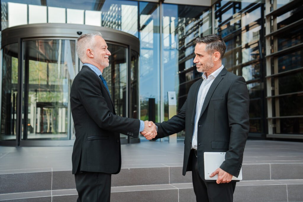 experienced senior executive warmly greeting younger business partner with confident handshake outside modern corporate building entrance, symbolizing successful mentorship, professional collaboration